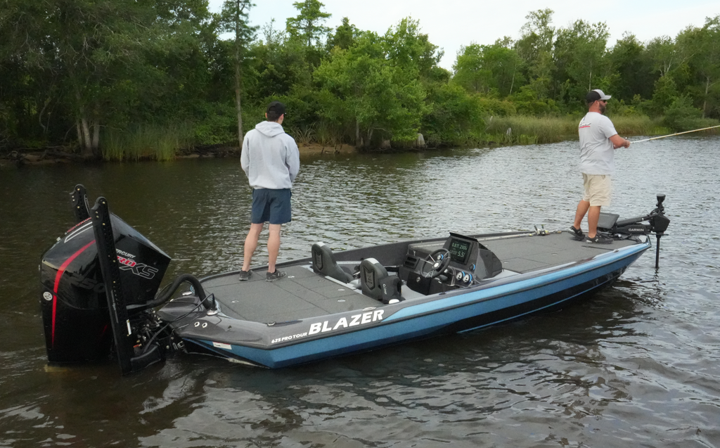 two men fishing from the bow and aft deck of the Blazer 625 Pro Tour. The boat is on a river, with a riverbank and forest in the background.
