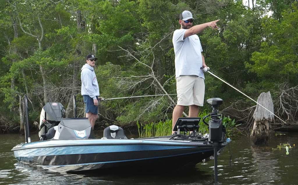 Two men fishing on a 625 Pro Tour. One man is pointing forward to the distance, while the other is standing with a fishing rod. The boat is on the water with the trolling motor pulling the boat along. In the background, you can see the riverbank and nearby forest.